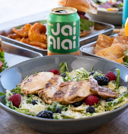 A salad with grilled chicken and berries, sandwiches, chips, and drinks, including a can labeled "Jai Alai" and a cocktail with an orange slice.