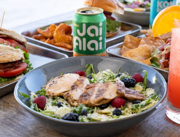A salad with grilled chicken and berries, sandwiches, chips, and drinks, including a can labeled "Jai Alai" and a cocktail with an orange slice.