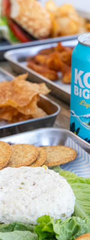 A picnic spread with sandwiches, a blue drink, chips, and fresh greens on a checkered blanket.
