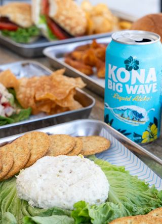 A spread of dishes includes a can of Kona Big Wave beer, burgers, chips, salad, and a platter with crackers and a spreadable dip.