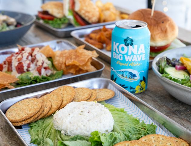 A spread of dishes includes a can of Kona Big Wave beer, burgers, chips, salad, and a platter with crackers and a spreadable dip.
