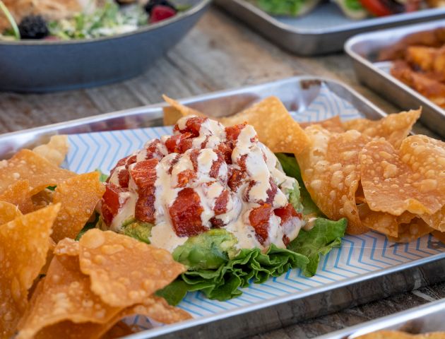A dish with crispy chips, lettuce, diced tomatoes, and a creamy sauce drizzle is presented on a metal tray.