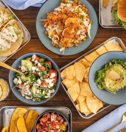 A spread of grilled chicken, fries, tacos, salads, hummus, pita chips, and a drink on a wooden table.