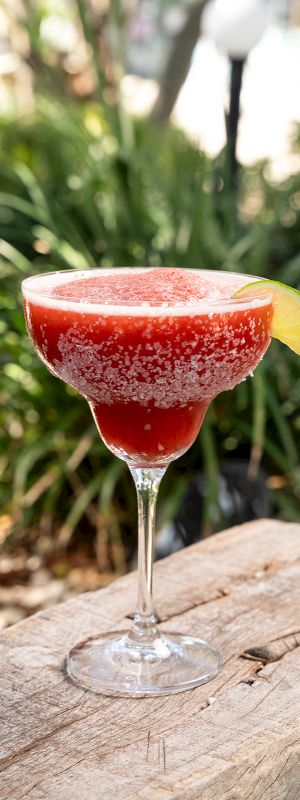 A red cocktail in a martini glass garnished with a pineapple slice and a cherry, set outdoors on rocks with greenery in the background.