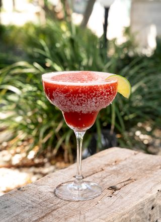 A red frozen cocktail in a martini glass with a lime wedge sits on a wooden surface outdoors.