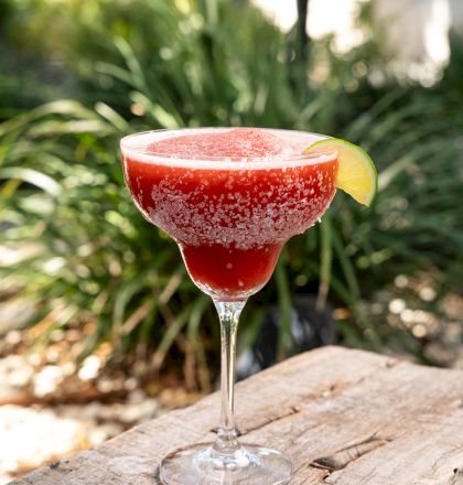 A salt-rimmed red margarita cocktail with lime on top sits on a wooden table, outdoors with greenery in soft focus behind it.