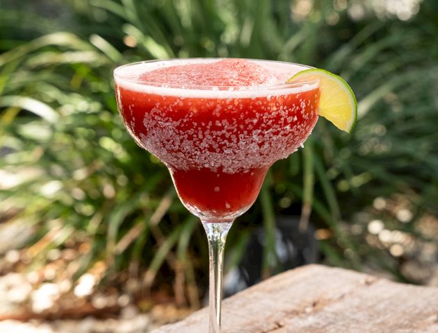 A red frozen cocktail in a martini glass with a lime wedge sits on a wooden surface outdoors.