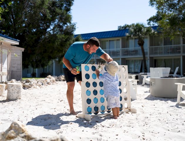 A person and a child play a giant Connect Four game on a sandy beach, with buildings and trees in the background.