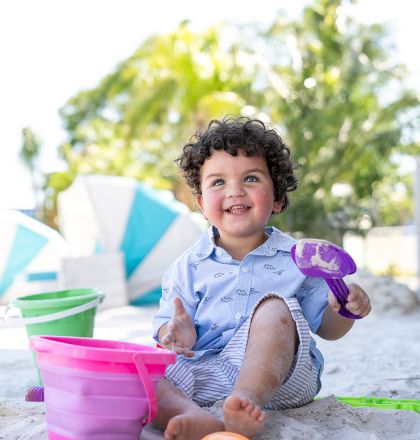A child in a blue shirt plays with a bucket and spade on a sandy beach, surrounded by trees and umbrellas in the background.