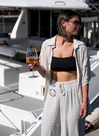 A person in a striped outfit holds a glass of drink, standing on a boat with sunglasses on, enjoying the sun.