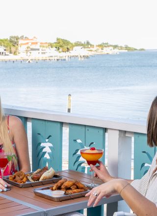 Two people enjoying drinks and food at an outdoor table by the water, with a scenic view in the background.