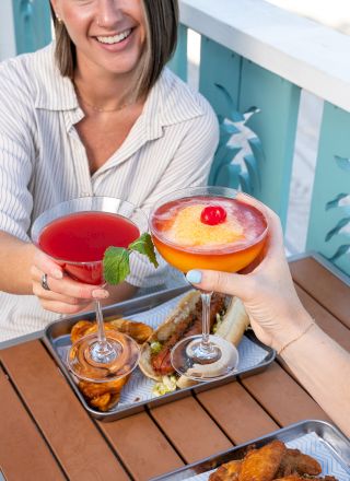 Two people toast with cocktails over a table with trays of food, including wings and fries. They smile by a blue railing with a pineapple design.