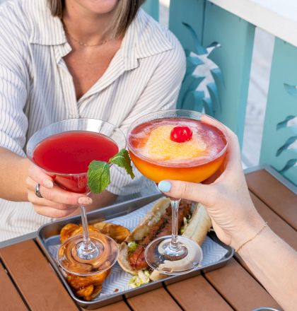 Two people toast with cocktails over a table with trays of food, including wings and fries. They smile by a blue railing with a pineapple design.