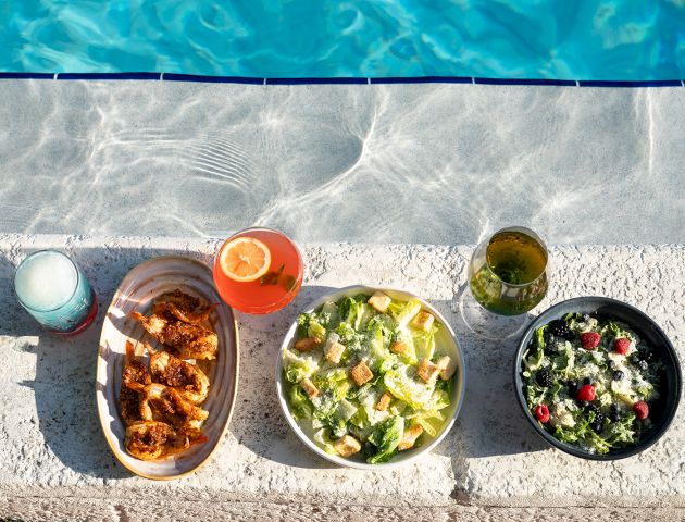 Plates of chicken, salad, drinks, and more are placed beside a pool, creating a refreshing summer meal setting.
