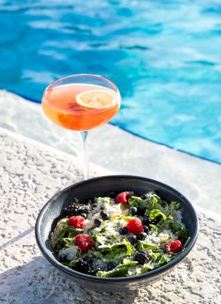 A bowl of salad with cherry tomatoes and a cocktail with a lemon slice by the poolside, creating a refreshing, summery vibe.