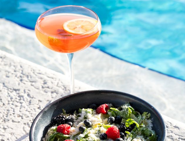 A bowl of salad with cherry tomatoes and a cocktail with a lemon slice by the poolside, creating a refreshing, summery vibe.