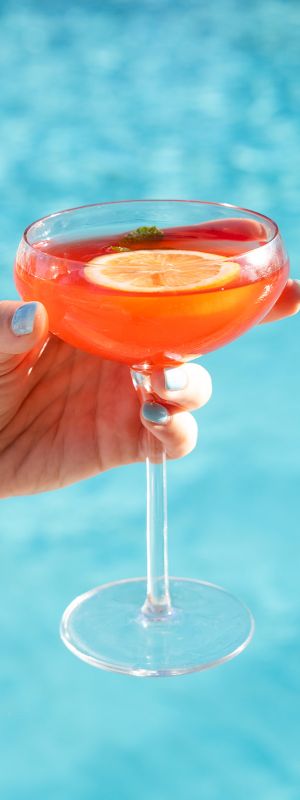 A hand squeezing a red, circular object with water streaming from it into a pool, likely a squeeze toy or bottle.