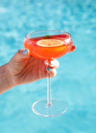 A hand holding a cocktail glass with a vibrant orange drink garnished with a lime slice by a pool.