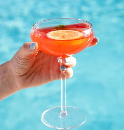 A hand squeezing a red, circular object with water streaming from it into a pool, likely a squeeze toy or bottle.