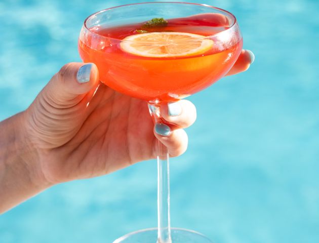 A hand holding a cocktail glass with a vibrant orange drink garnished with a lime slice by a pool.