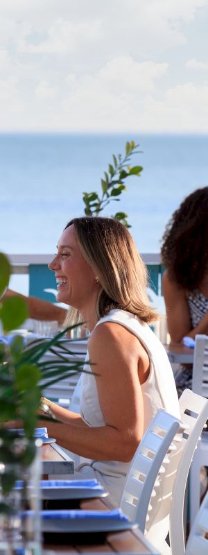 A group dines at an outdoor seaside restaurant, drinks and greenery on the table, blue ocean beyond, a waiter stands nearby.