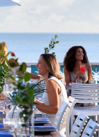 A group dines at an outdoor seaside restaurant, drinks and greenery on the table, blue ocean beyond, a waiter stands nearby.