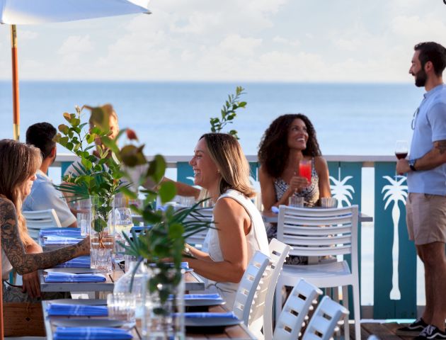 A group dines at an outdoor seaside restaurant, drinks and greenery on the table, blue ocean beyond, a waiter stands nearby.