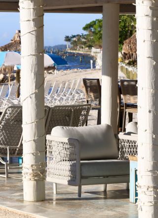Outdoor lounge area with wicker furniture under a pergola, overlooking a beach with blue and white striped umbrellas.