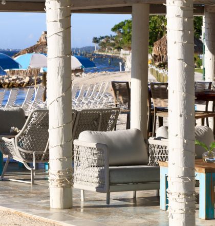 Outdoor lounge area with wicker furniture under a pergola, overlooking a beach with blue and white striped umbrellas.