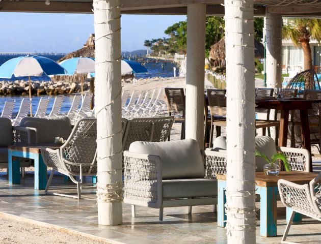 Outdoor lounge area with wicker furniture under a pergola, overlooking a beach with blue and white striped umbrellas.