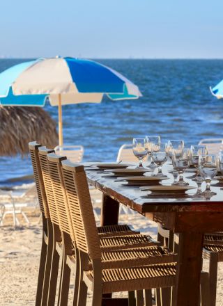 A beachside setup with a dining table and chairs, nearby umbrellas and loungers, facing the ocean under a clear blue sky.