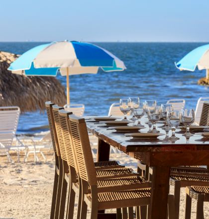 A sunny beach scene with a sandy patio, wooden tables and chairs, striped umbrellas, and a clear blue sky behind seaside buildings.