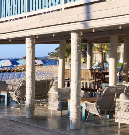Outdoor seating area under a beachfront structure with wicker chairs and tables, overlooking an ocean with umbrellas and lounge chairs.