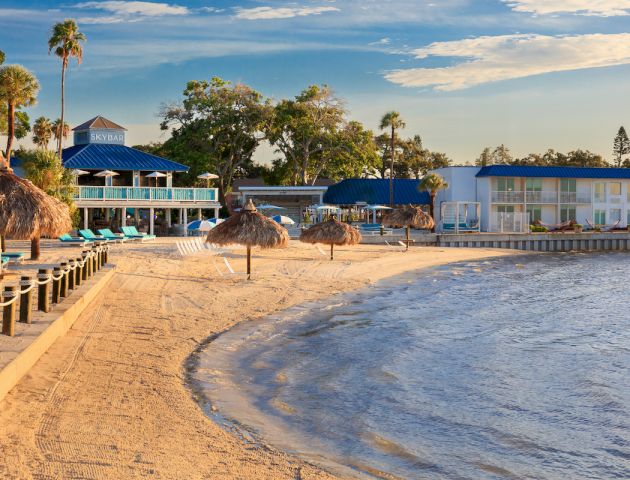 A sandy beach with palm trees, thatched umbrellas, and a resort with blue roofs under a clear sky. Lounge chairs line the shore.