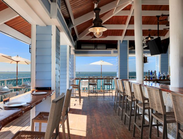 An open-air beachside bar with wooden stools, tables, and a view of the ocean under a clear blue sky with umbrellas outside.