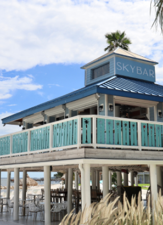 A beachside bar with a turquoise railing and sign reading "SkyBar," set against a backdrop of palm trees and a partly cloudy sky.
