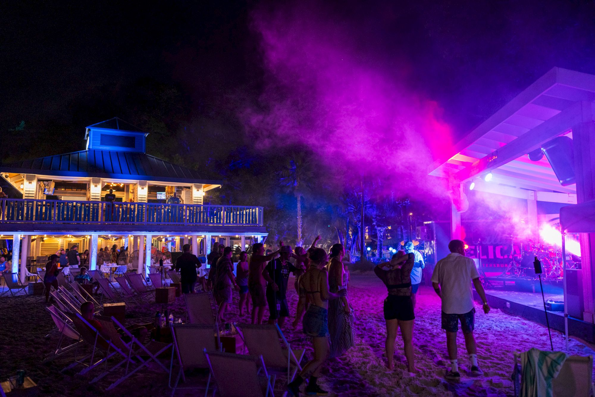 A beach party at night with people dancing near a stage and a lit building in the background, under colorful lights and smoke.