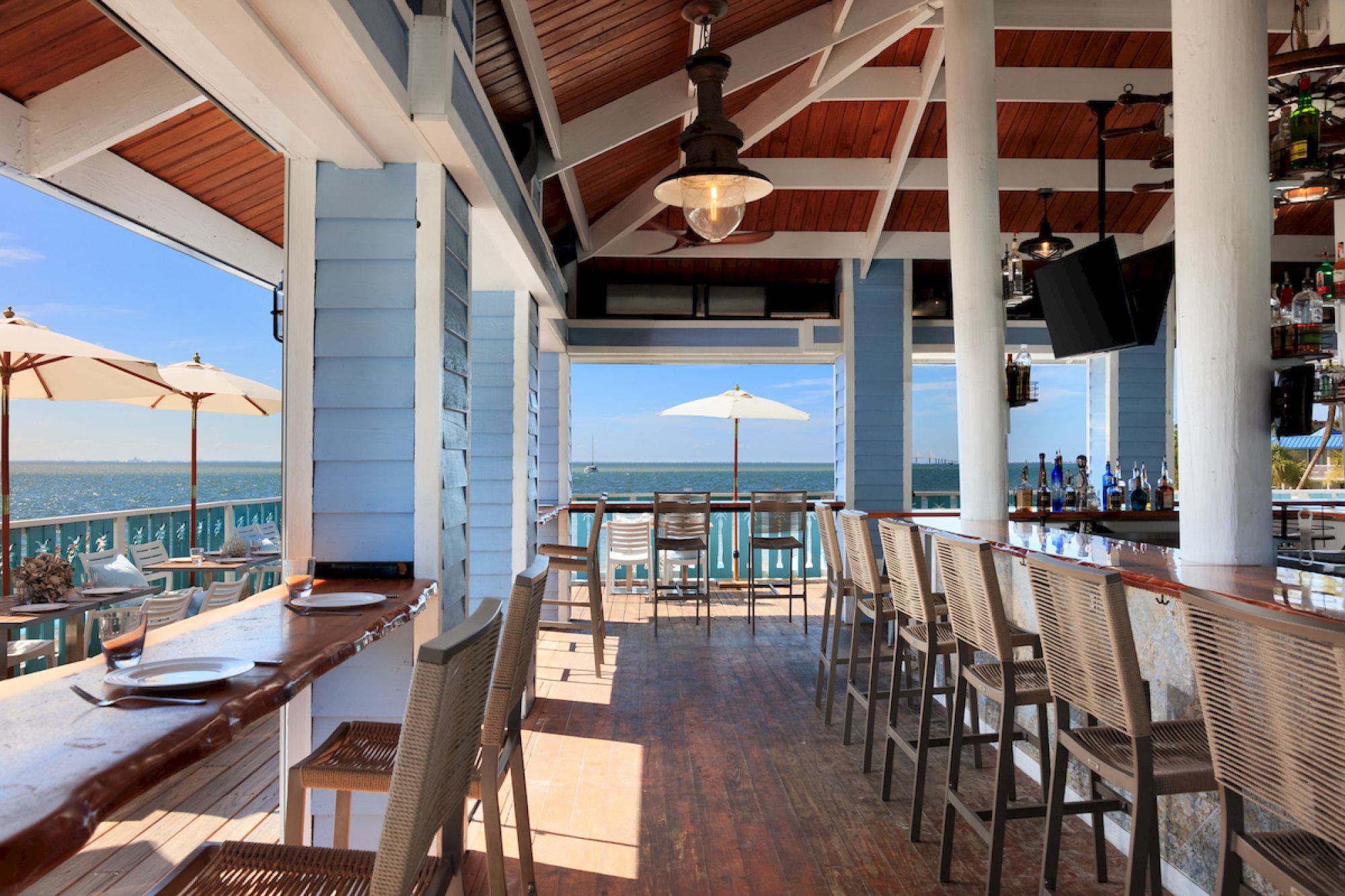An open-air beachfront bar with stools, tables, and umbrellas; ocean view in the background.