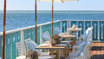 A sunny seaside patio with turquoise railing, white chairs, wooden tables set with plates and shells, and umbrellas over-looking the ocean.