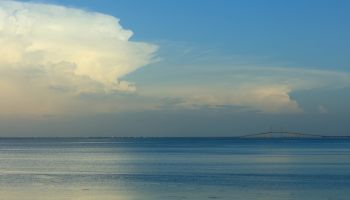 A calm seascape at dusk with a smooth blue-gray ocean, soft clouds on the horizon, and a hint of a distant landmass or bridge to the right, serene and wide.