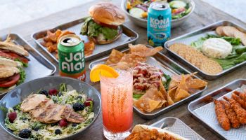 An array of food including burgers, salads, chips, sandwiches, with drinks like a pink beverage and canned drinks, displayed on trays.