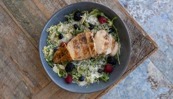 A dish with sliced grilled chicken on a bed of greens, berries, and shaved parmesan, served in a gray bowl on a wooden table.