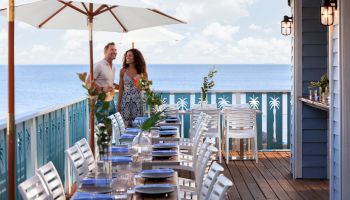 Two people stroll on a seaside restaurant deck with white chairs, tables set for dining, umbrellas, and a blue ocean in the background.