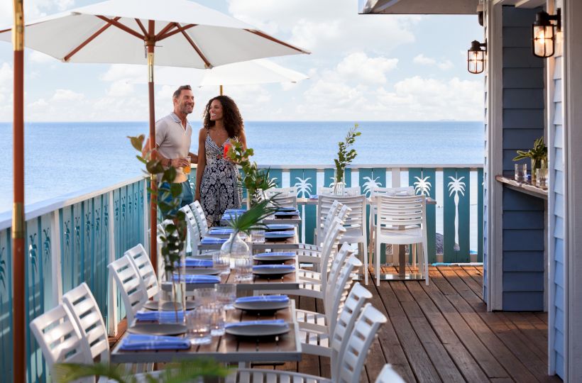 Two people stroll on a seaside restaurant deck with white chairs, tables set for dining, umbrellas, and a blue ocean in the background.