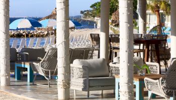 A beachside lounge with woven chairs, white columns, and blue tables under a shaded patio area, overviewing sand and blue umbrellas.