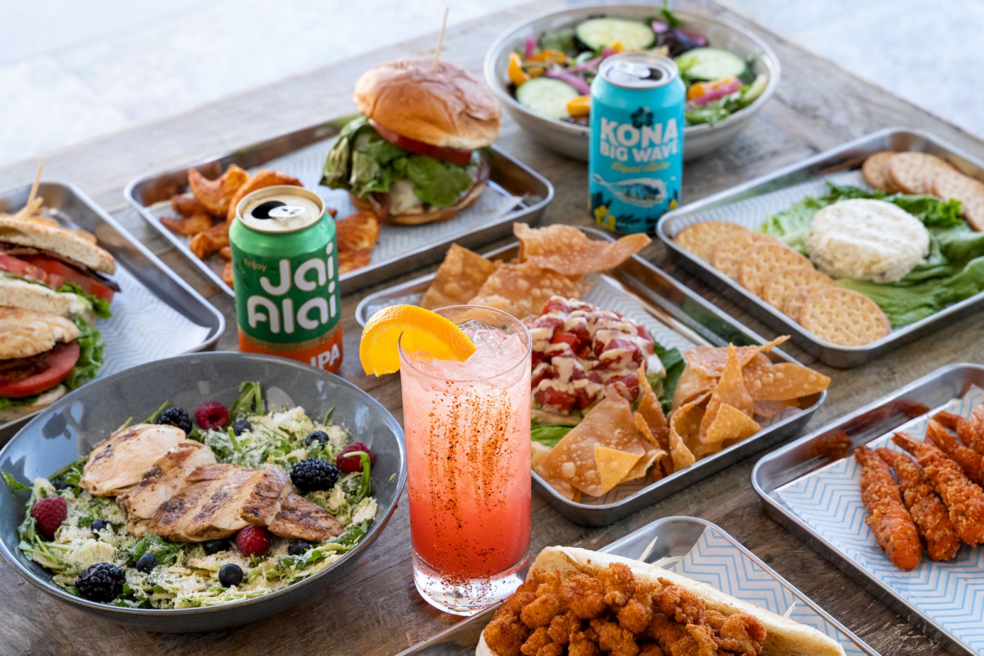 An array of dishes including salads, sandwiches, chips, drinks, and fried foods on trays, alongside a pink beverage with an orange slice.