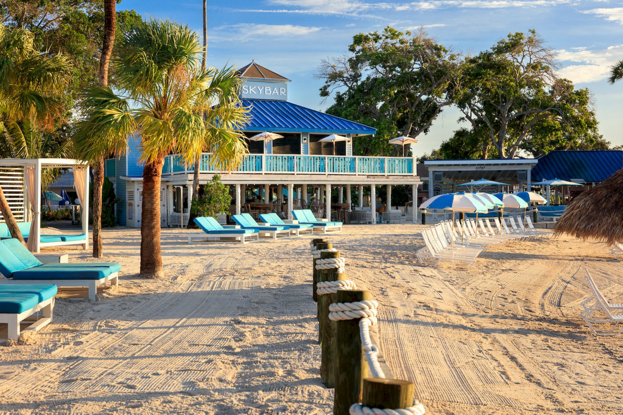 A sandy beach scene with lounge chairs, palm trees, and a building marked "SKYBAR" in the background under a clear blue sky.
