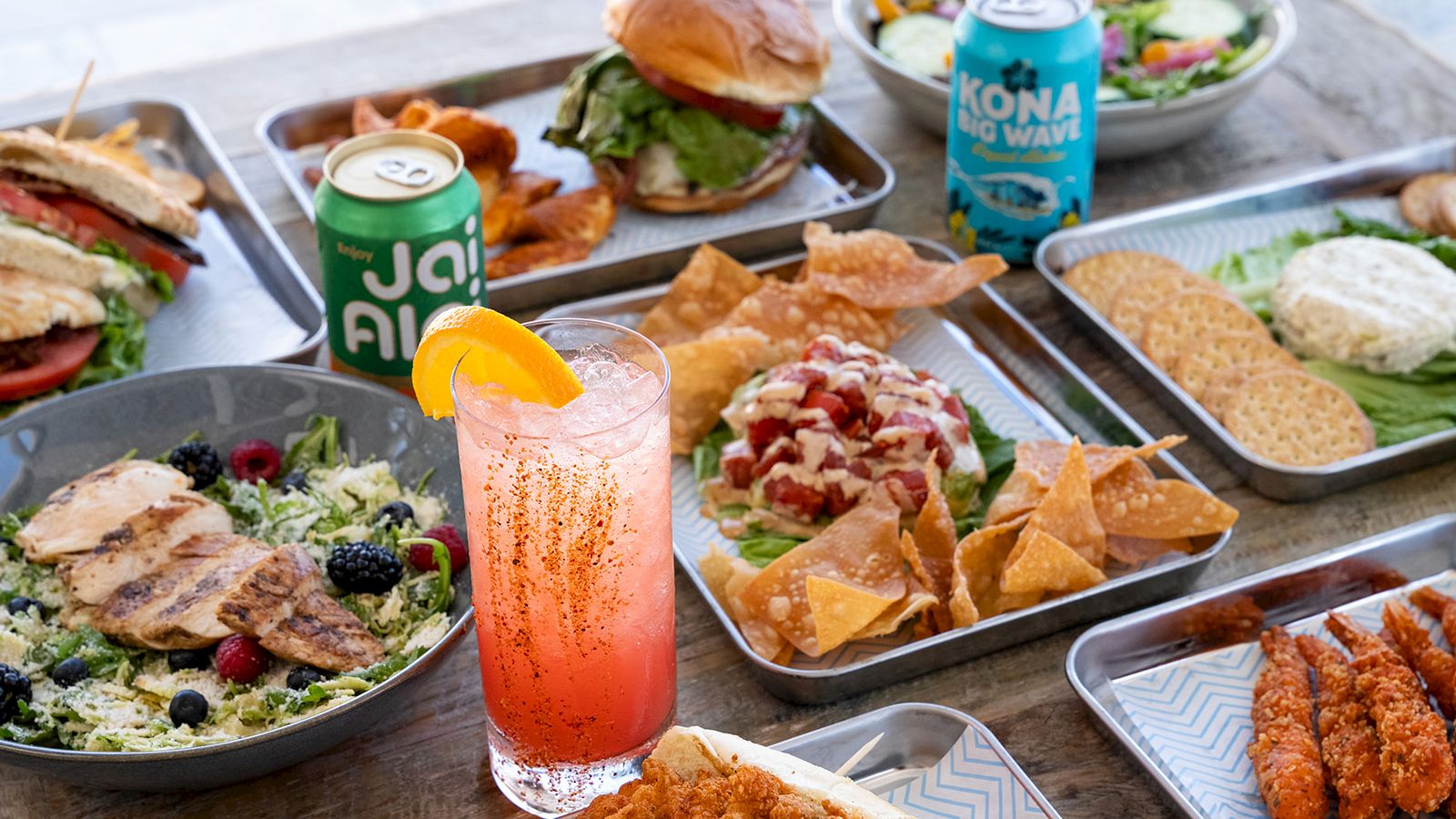 A vibrant spread of artisan tacos, nachos, grilled dishes, salads, fried bites, and two canned drinks plus a pink citrus beverage on a wooden table.