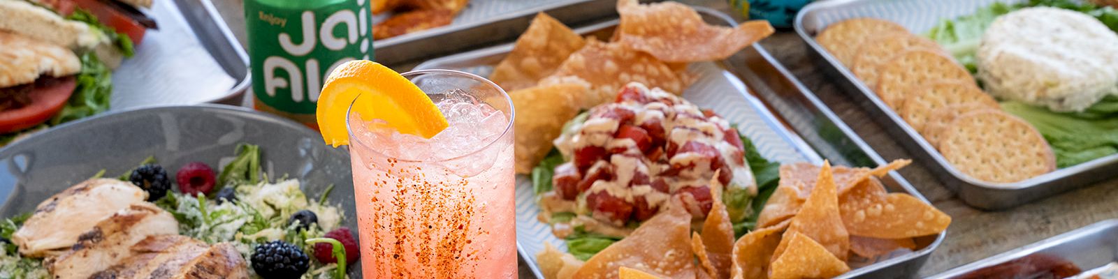 A vibrant spread of artisan tacos, nachos, grilled dishes, salads, fried bites, and two canned drinks plus a pink citrus beverage on a wooden table.