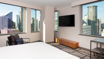 A modern room with a bed, chair, wall-mounted TV, and city views through large windows, featuring skyscrapers outside.